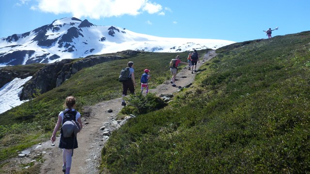Multiple hikers of different ages walk up a trail with a snow-covered mountain beyond.