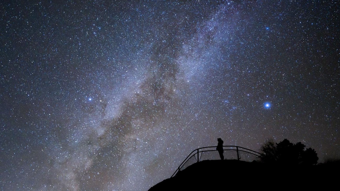 The black silhouette of a person standing at a viewpoint with the milky way in the backgroundn