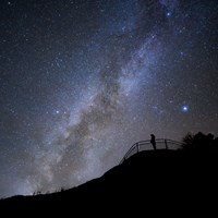 A silhouetted figure stands at a fenced overlook with the milky way arching above