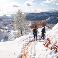 Two hikers walk on a snow covered trail with mountains in the background.