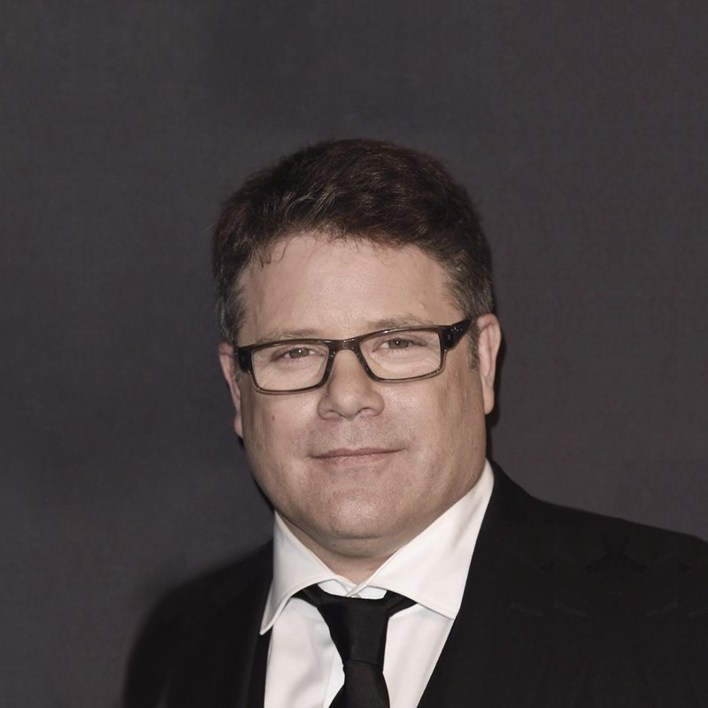 Image of Sean Astin wearing a black suit coat, white shirt, black tie, and glasses.