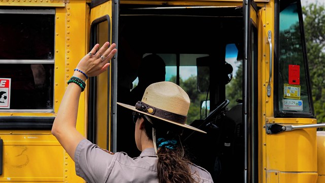 A Park Ranger waves at students on a bright yellow school bus.
