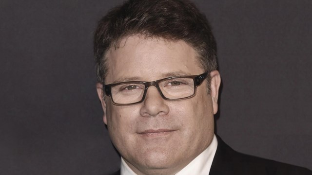 Image of Sean Astin wearing a black suit coat, white shirt, black tie, and glasses.