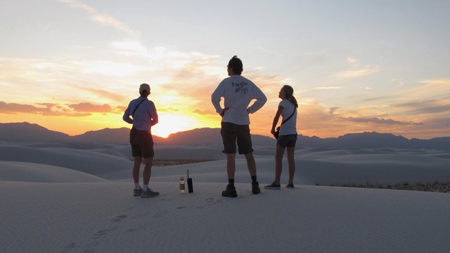three people stand on a white sand dune silhouetted by sunset over the mountains.