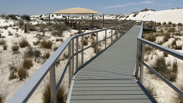 a boardwalk extends into the distance among white sands dunes. A shade paviliion can be seen ahead.
