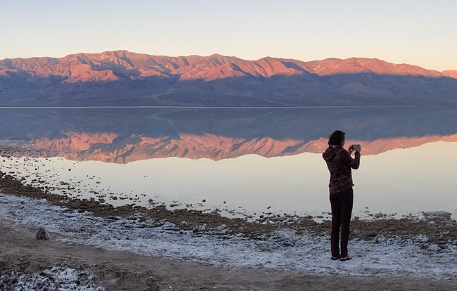 A person stands with back to camera holding a camera. A large body of water reflects pink mountains.