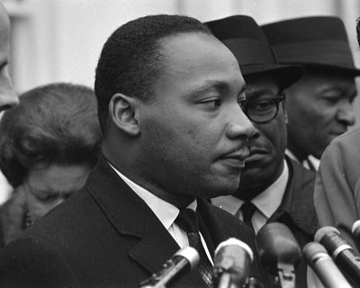 Martin Luther King Jr. standing behind a series of microphones and surrounded by a crowd.