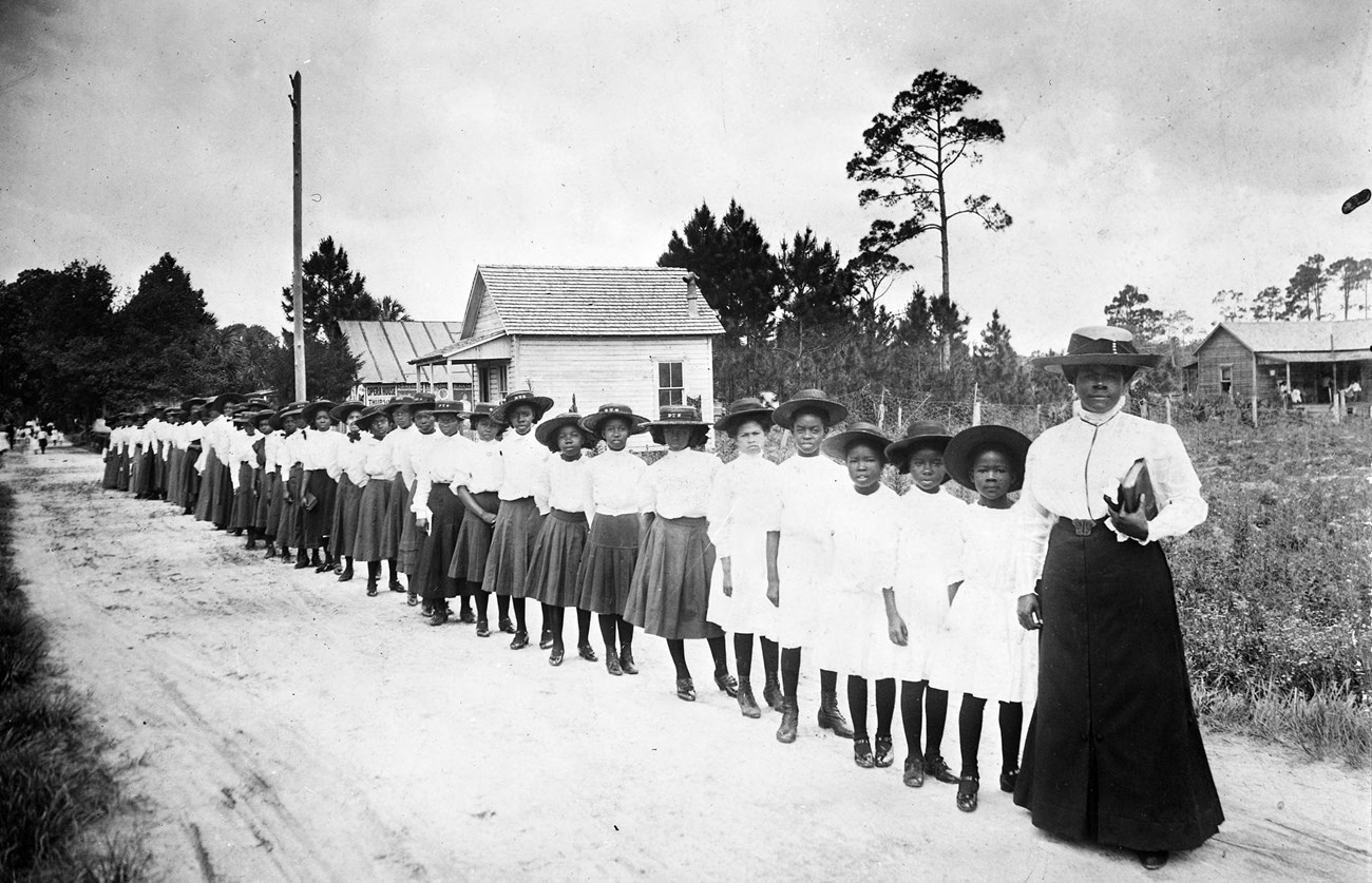 Mary McLeod Bethune with a line of girls standing on a road in front of a wood school house.