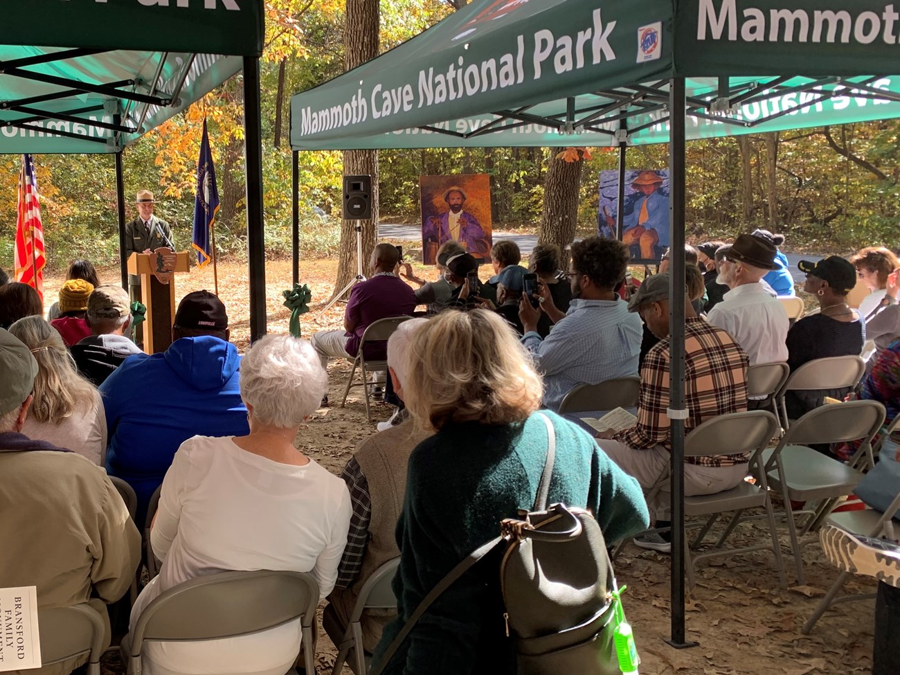 A large group of people sit under green canopies that say "Mammoth Cave National Park" while listening to a man speak at a podium.