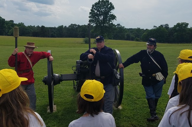 Three volunteers in living history uniforms flank a cannon while students ask questions in front of them.