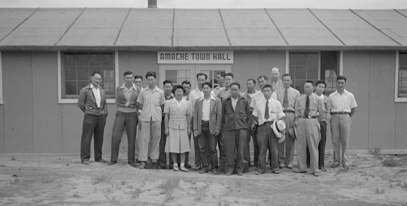 Black and white photograph of a group of Japanese Americans standing in front of a barrack structure.