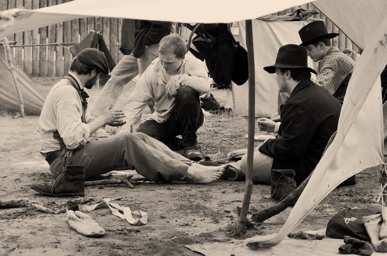 Four men in ragged Civil War era clothes sit on the ground under a makeshift shelter.
