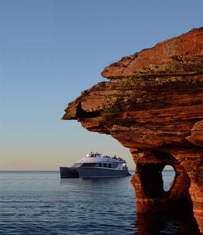 A white and grey catamaran with passengers is viewed next to a red sandstone arch.