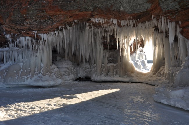Ice formations frame an opening in a rocky cliff near a frozen lake surface.