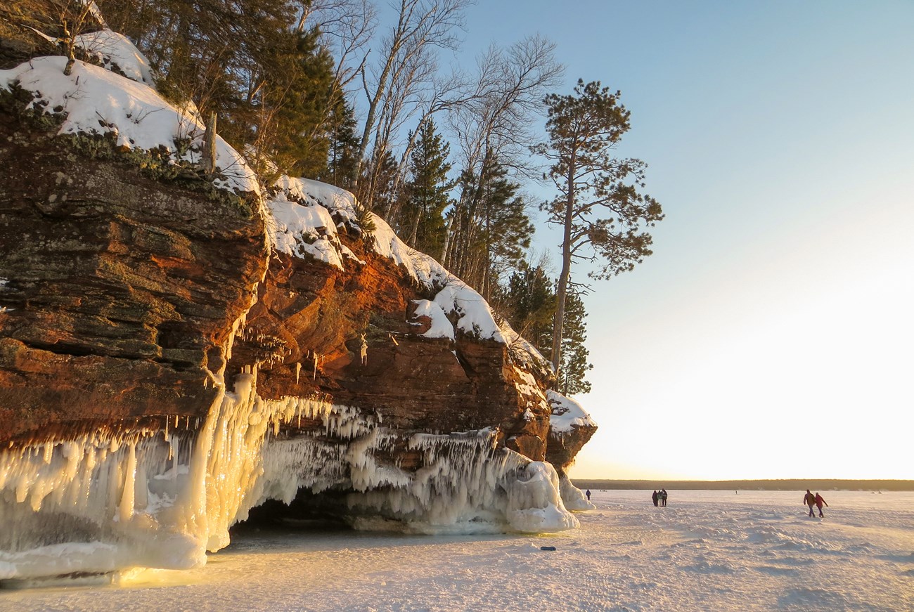 Mainland Ice Caves at Apostle Islands National Lakeshore in 2014 Photograph of sandstone cliffs draped in icicles and people walking across a frozen lake.