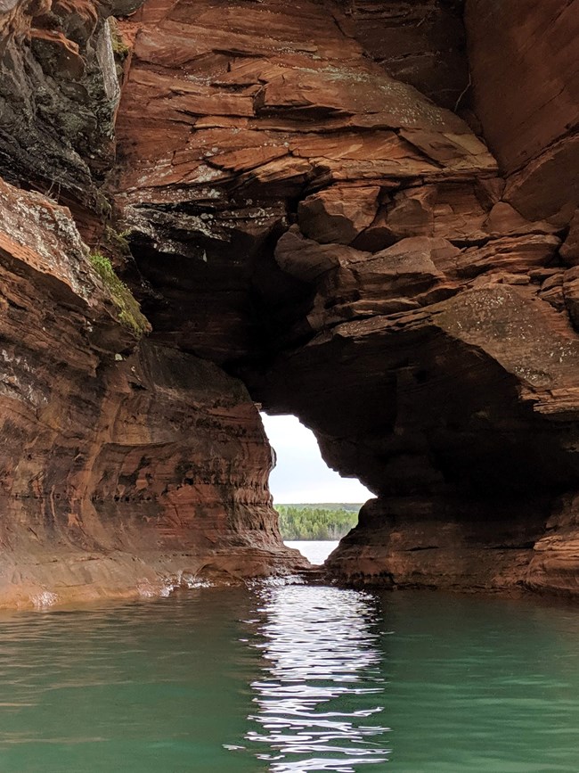 Hole in sandstone cliffs with green water in the foreground and distant trees across the water seen through the hole.