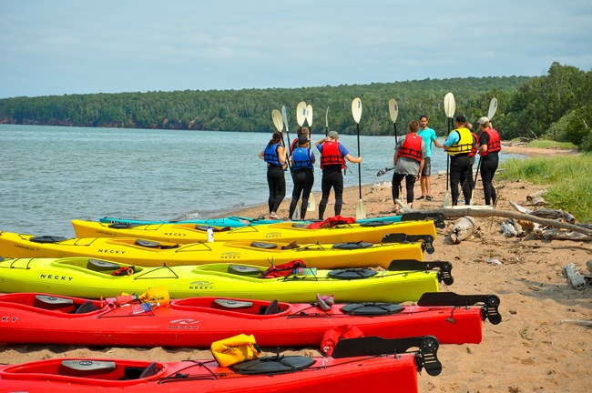 A group of kayaks resting on the shore and a people getting a safety talk before heading out to the sea caves.