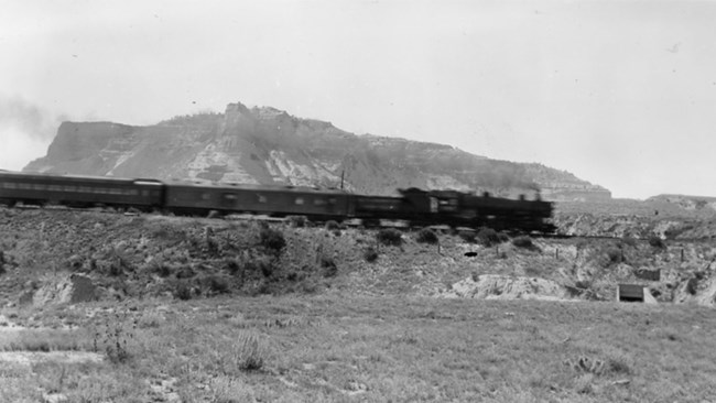 A steam engine makes its way past sandstone bluffs.