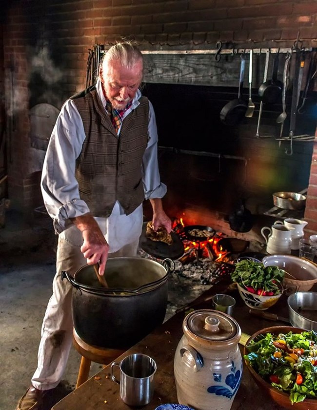 A man wearing 1840s style clothing cooks food in a large cast iron pot.