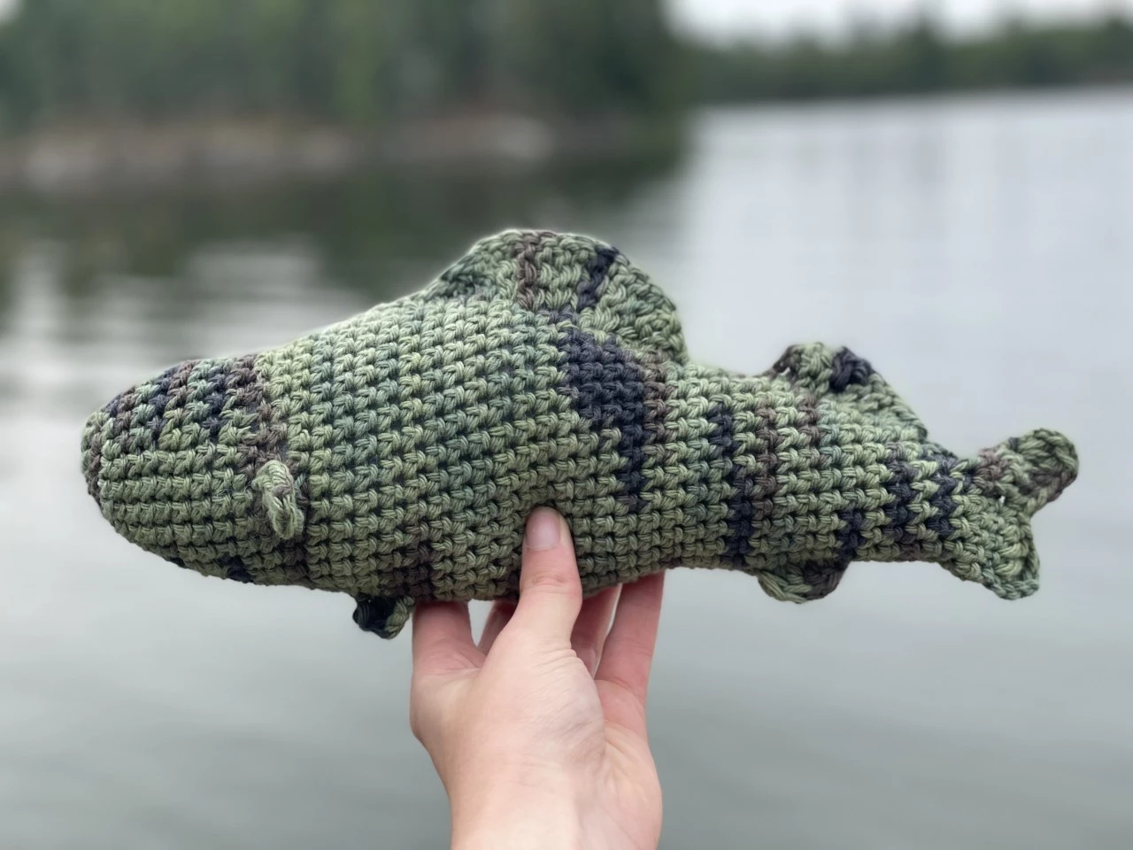 Crochet fish A crocheted fishing being held by a hand in front of a lake.