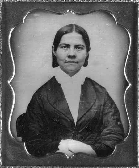 A middle-aged White woman, Lucy Stone, wearing a dark dress and white blouse, sitting and looking at the camera.