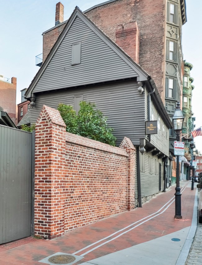 The Revere House is a two story wooden building painted gray. The high pitched roof is cedar shingle with a chimney on the right end of the house. The windows are panes of diamond glass. Shutters are only on the first floor.