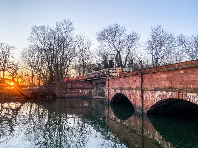 A red stone aqueduct spans a creek leading into a river, while the sun sets behind it.