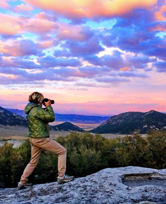 A photographer stands on a rock in a dramatic sunset vista with her camera raised.
