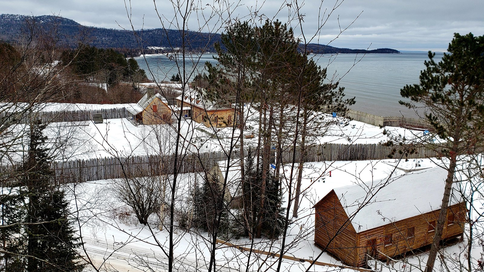 View from a mountain trail of wood buildings in the snow with a lake in the background.