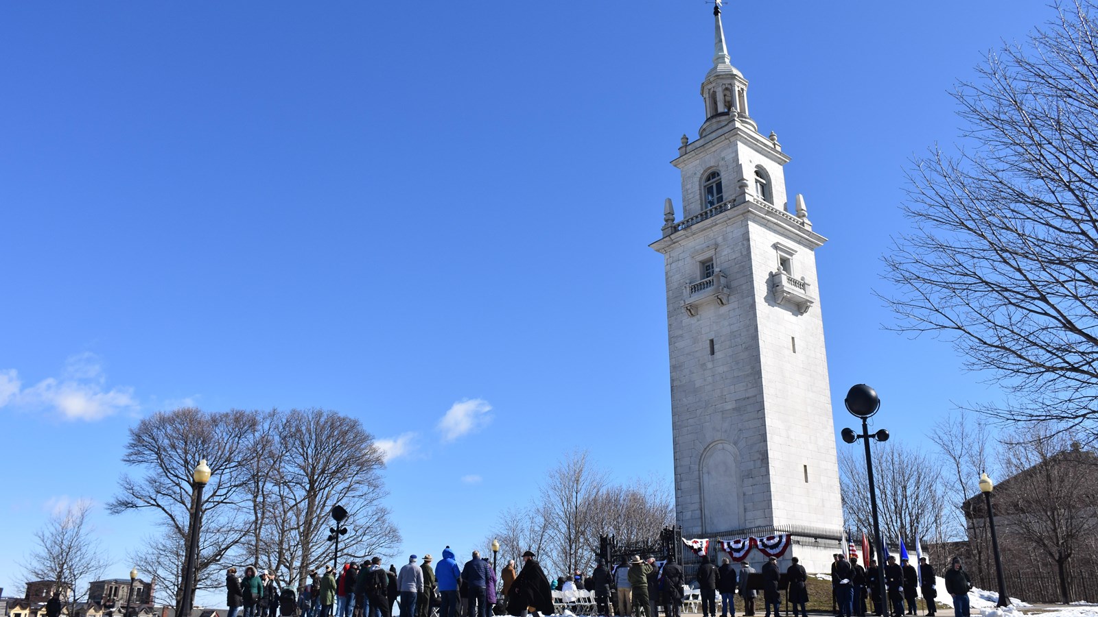 People standing around Dorchester Heights Monument. Trees are bare and ground has light snow.