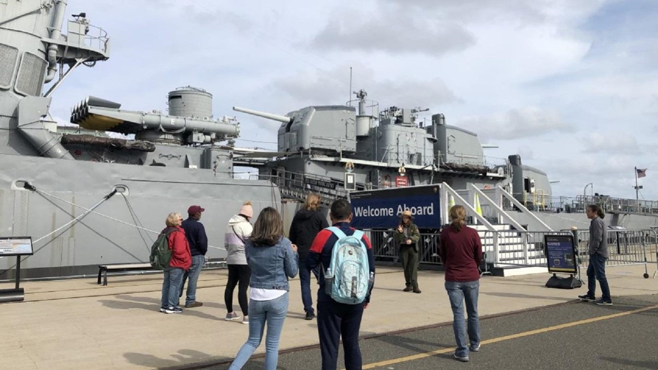 several small groups of people spread out looking at a ranger on the pier next to a destroyer.