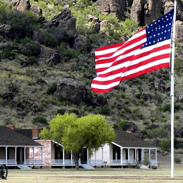 A large U.S. flag flies on a pole in front of several brick buildings