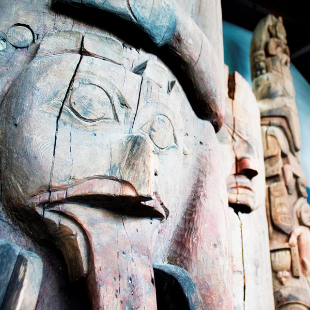 Close up of a face carved in old distressed wood; totem poles in the background.