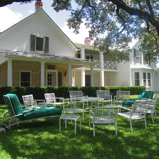 A circle of white chairs sit on the grass in front of a two-story, white-frame house. 