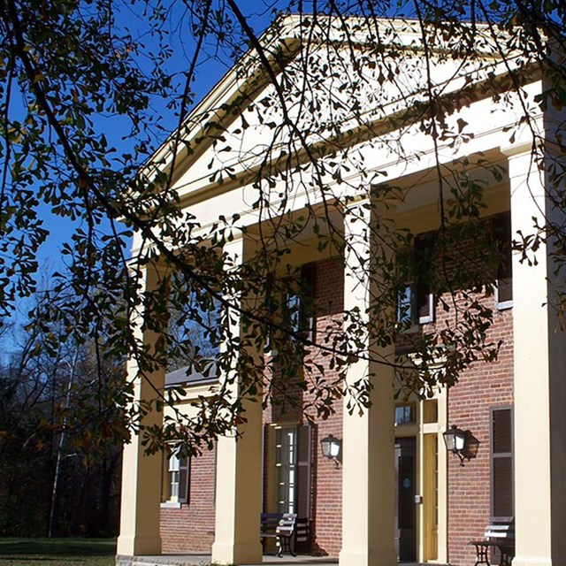 Two-story red brick building with four cream-colored support pillars on manicured lawn.