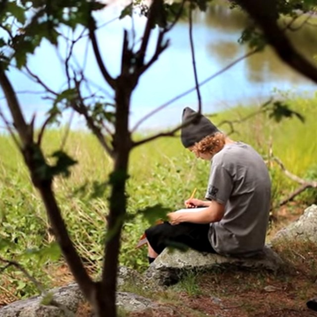 young man sitting on rock next to river writing in notebook