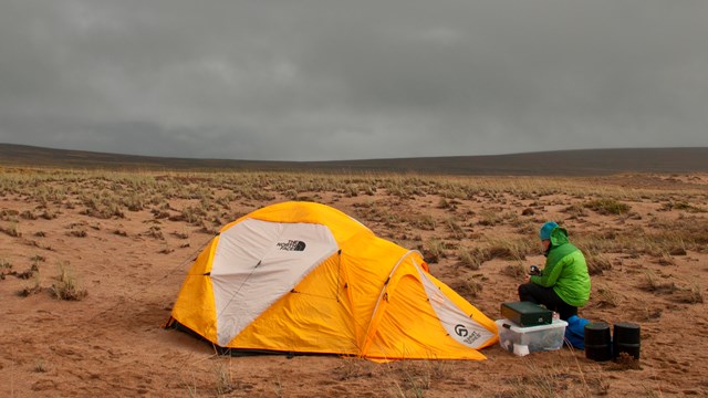 A yellow tent is pitched on bare ground as gray clouds loom above. 