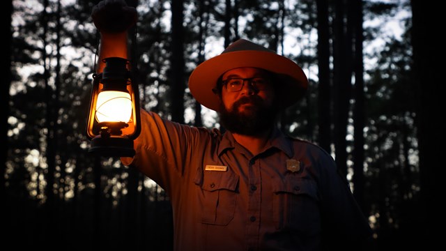 A male park ranger with glasses and beard holding a lit vintage lantern.