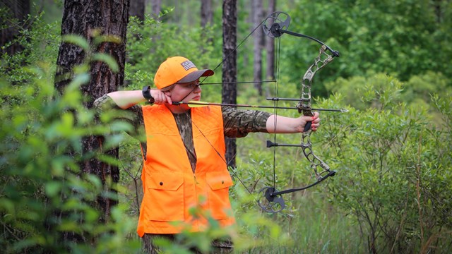 A woman wearing hunting orange vest and camo pattern clothing aiming a bow and arrow in the woods.