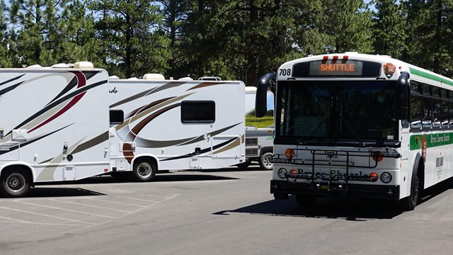 A shuttle drives behind 2 large RVs in parking spaces.