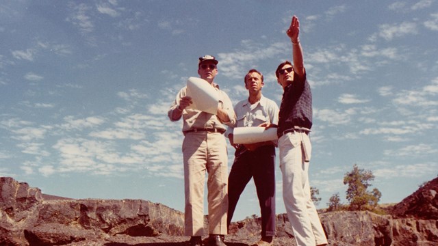 Three men look out on a lava landscape while holding papers. 