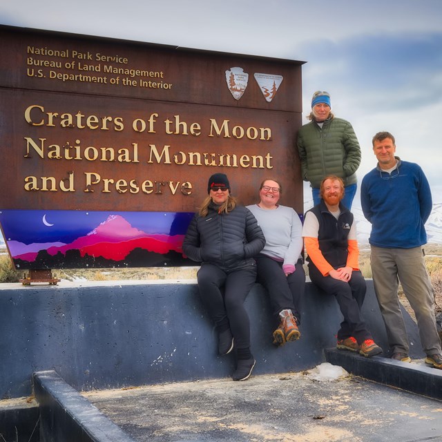 Group of 6 people sit and stand next to a metal sign that says Craters of the Moon National Monument