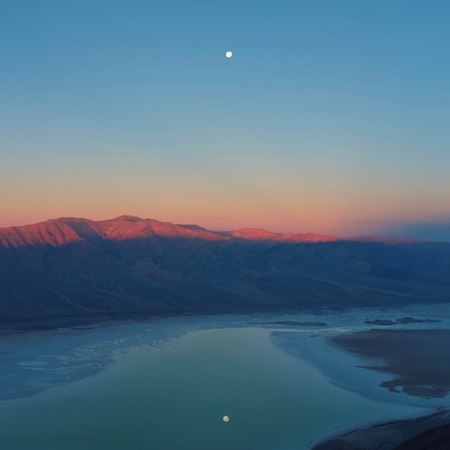 A sunset view over a valley filled with a shallow lake surrounded by desert mountains.