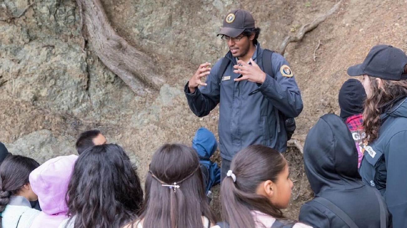 A young man in a volunteer uniform speaks to a group of kids outside.