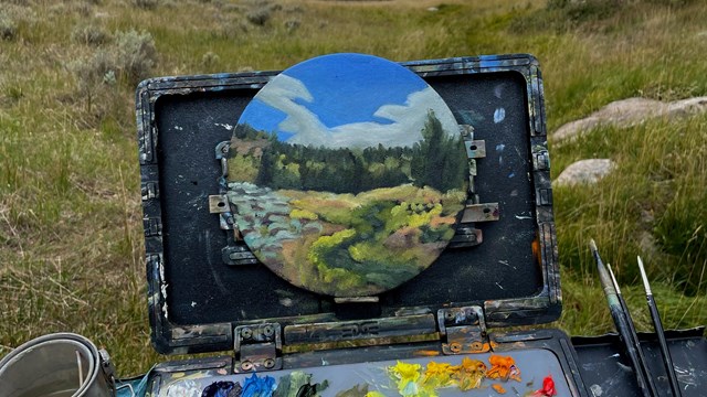 A photo of a painter's palette and small painting in front of a meadow