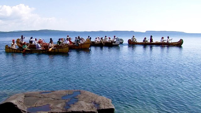 Birch canoes in a bay paddled by people in colorful historic clothing.