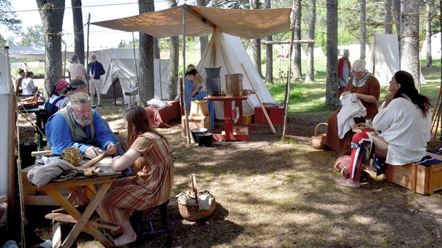 People wearing historic fur trade era clothing, seated under white canvas tents.
