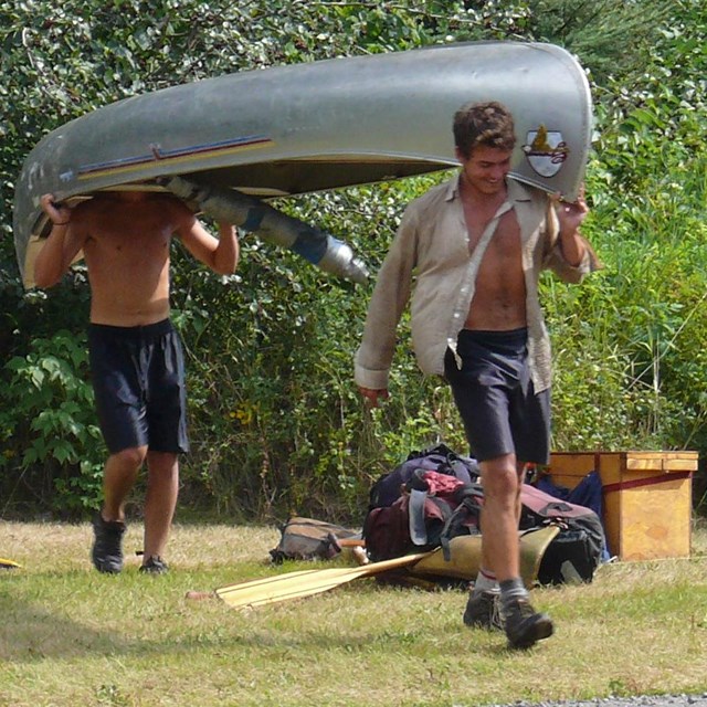Two people carrying an aluminum canoe with other gear strewn on the ground.