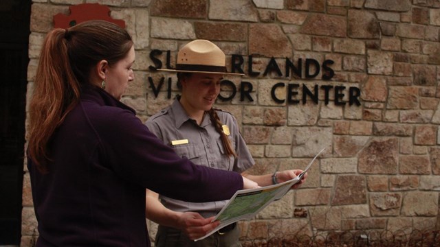A ranger standing outside a visitor center helping a visitor by showing them a map.