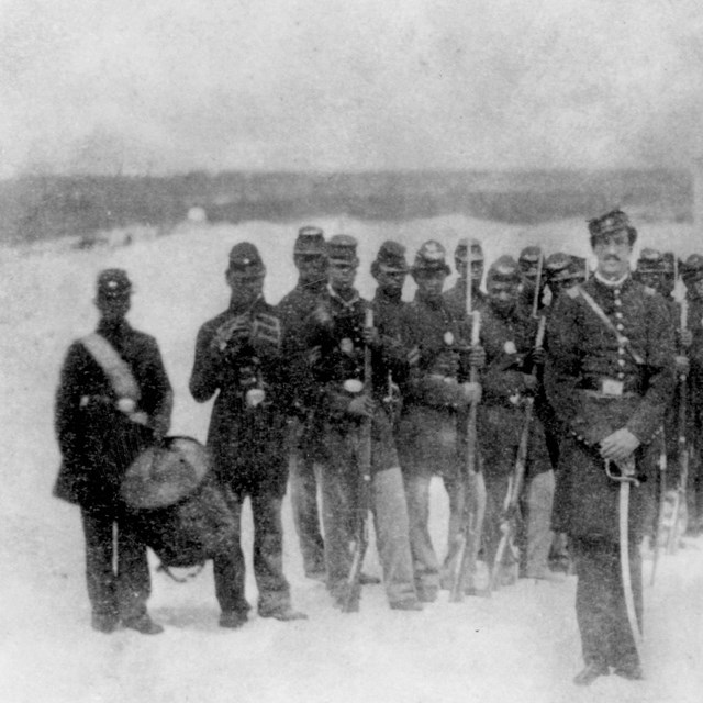 Black and white photo of African American soldiers formed on a beach.